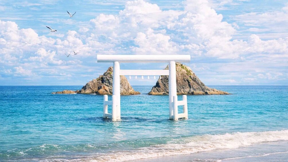 A white torii gate in the ocean with rocky formations in the background. 1