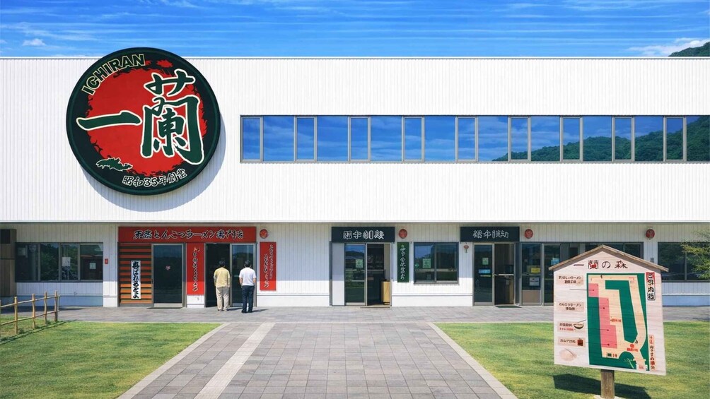A modern building with a large circular sign featuring Japanese characters. There are two people standing in front of the entrance, and a map sign is visible to the right. 3