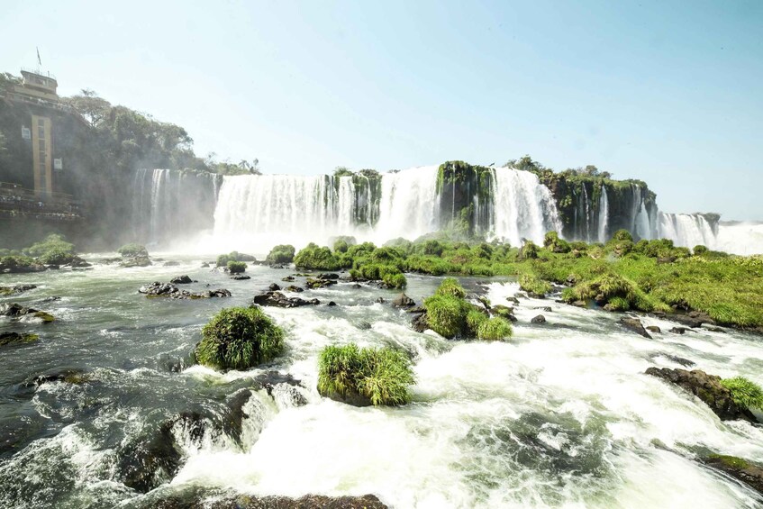 A multi-tiered waterfall with a river in the foreground, surrounded by lush greenery and a building on the left. 1
