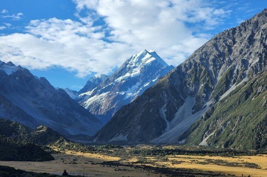 A mountain range with a prominent snow-capped peak, a valley with sparse vegetation, and a clear sky. 1