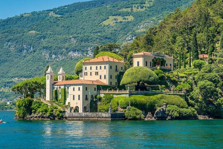 A historic villa with a tower, surrounded by lush greenery and a lake. 1