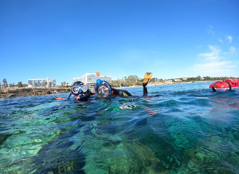 Two snorkelers in clear blue water with a rocky shoreline and buildings in the background. 1
