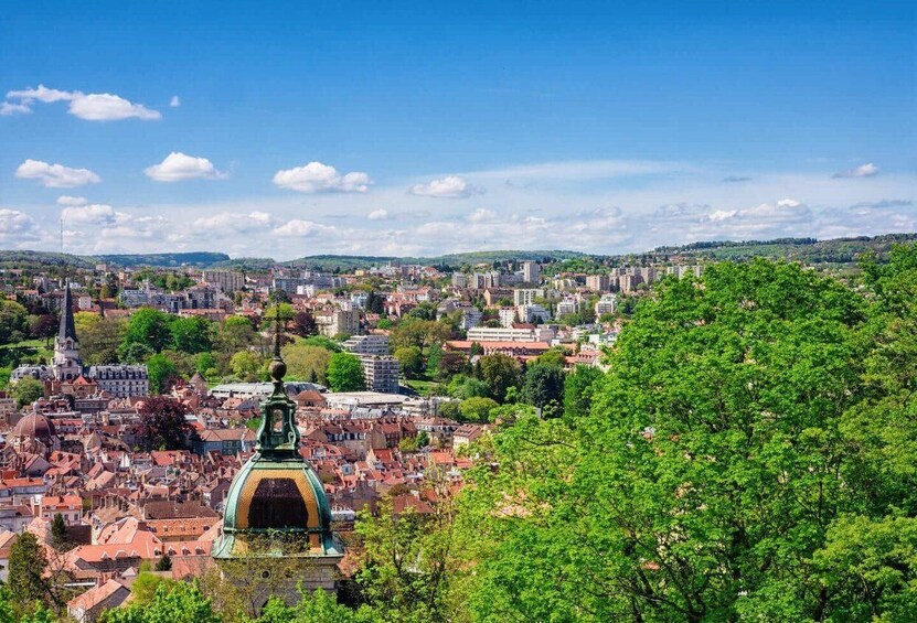 A panoramic view of a city with a prominent green-domed building in the foreground, surrounded by red-roofed buildings and a clear blue sky. 2