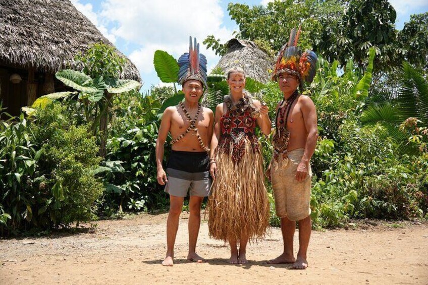Three people in traditional attire standing in a tropical setting with a thatched-roof hut and lush greenery in the background. 1