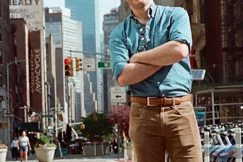 A person standing with arms crossed on a city street with tall buildings and traffic lights in the background. 3