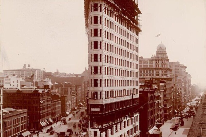 A historical cityscape with a prominent building under construction, a large dome structure, and a busy street with pedestrians and vehicles. 2