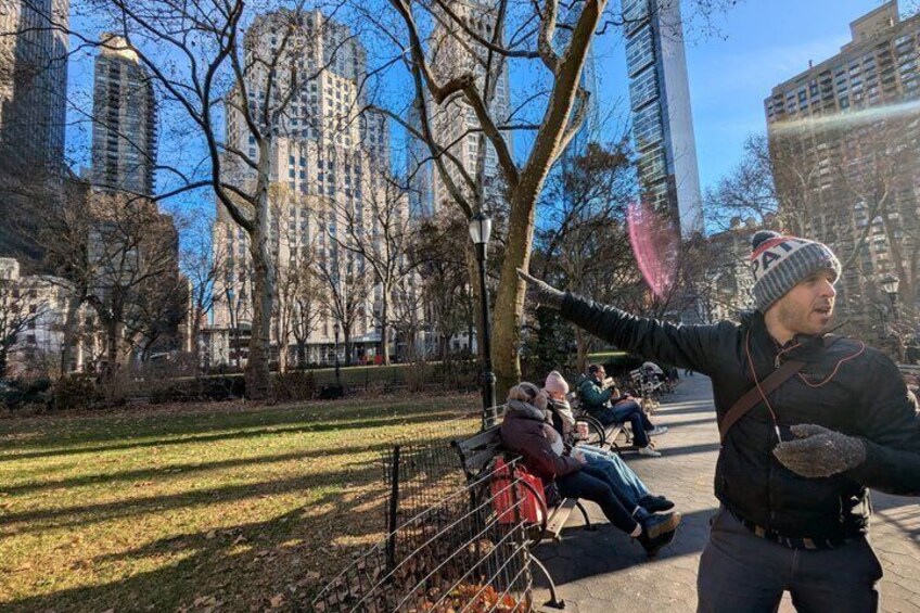 A park with people sitting on benches, trees, and tall buildings in the background. 1