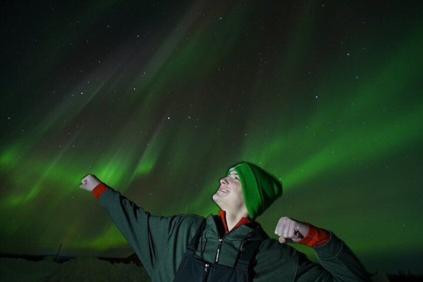 A person wearing a green hat and jacket, pointing towards the sky with green aurora borealis. 2