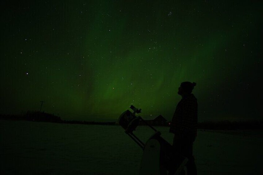 A person with a telescope on a snowy landscape under a green aurora. 1