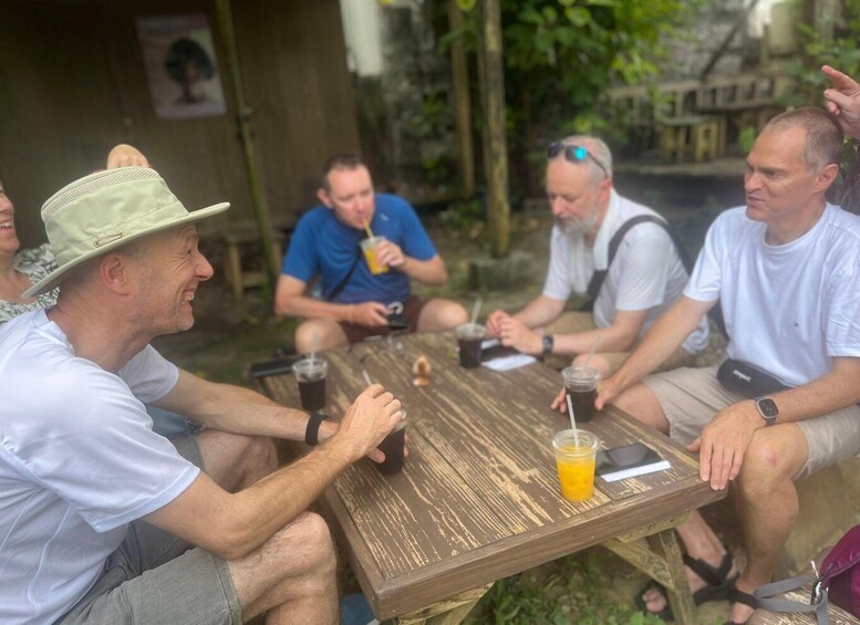 A group of people sitting around a wooden table outdoors, engaged in conversation. 1