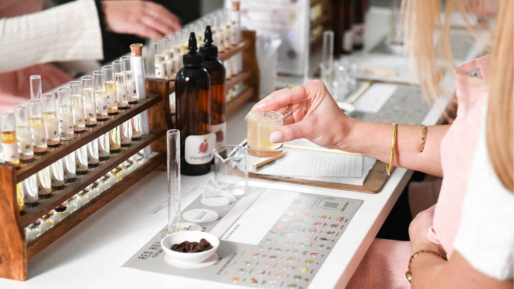 A person holding a small glass of liquid, with a variety of bottles and test tubes on a table. 3