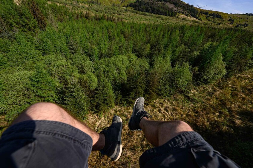 A person's legs and feet, wearing black shorts and sneakers, sitting on a grassy hill with a forested valley in the background. 4