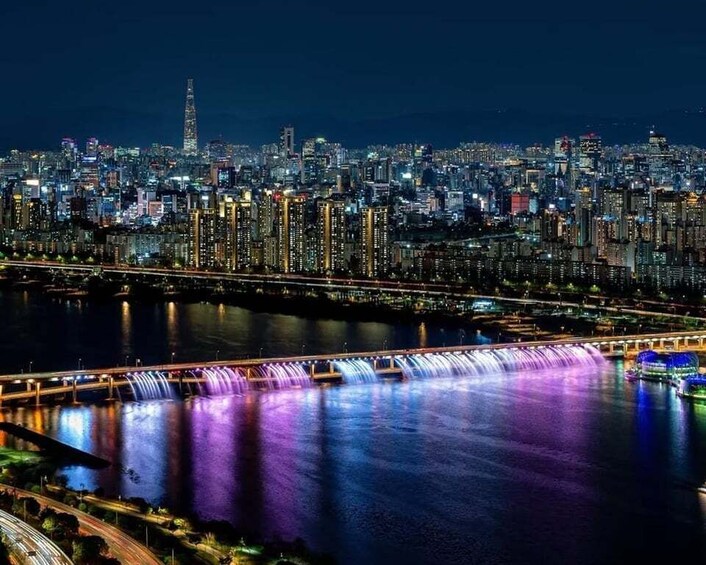 A nighttime cityscape with illuminated buildings, a river, and a bridge with water features. 3