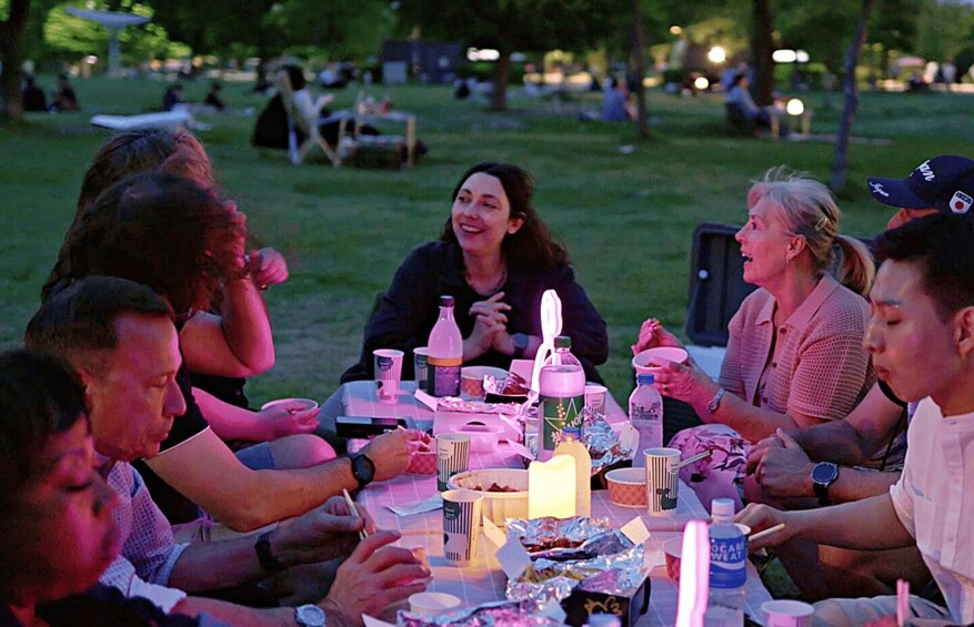 A group of people sitting around a table with food and drinks, outdoors in a park. 4