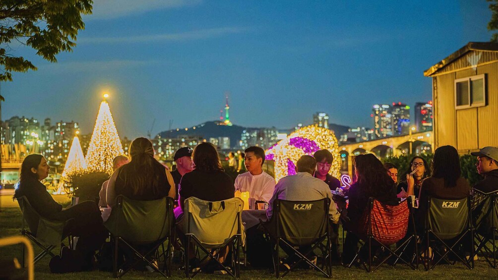 A group of people seated at an outdoor event with illuminated decorations and a cityscape in the background. 2