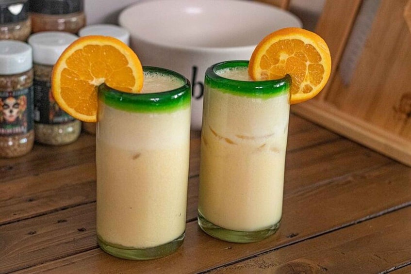Two glasses of a creamy beverage with a slice of orange garnish, placed on a wooden surface with spices in the background. 2