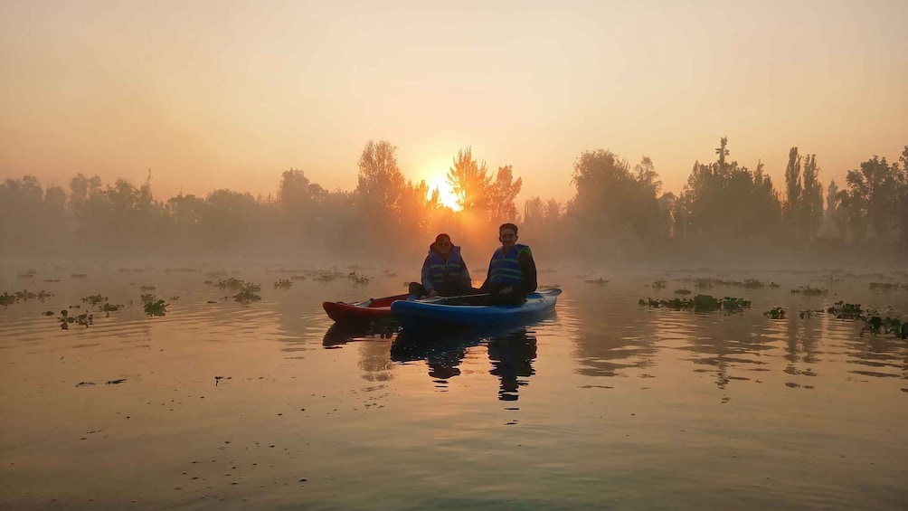 Two people in a blue kayak on a calm body of water with a sunrise in the background. 1