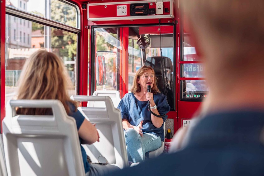 A red bus interior with a person holding a microphone, seated passengers, and a fire extinguisher mounted on the wall. 2