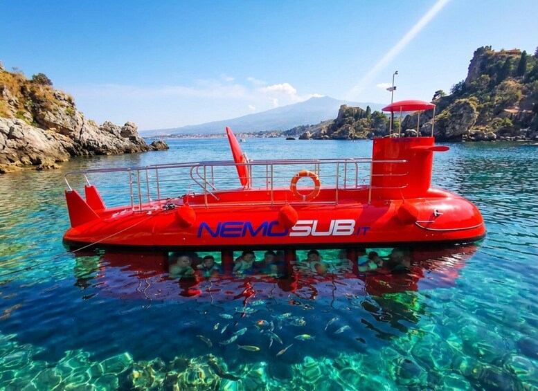 A red inflatable boat named NEMC SUB.it floating on clear blue water with rocky cliffs and a mountain in the background. 1