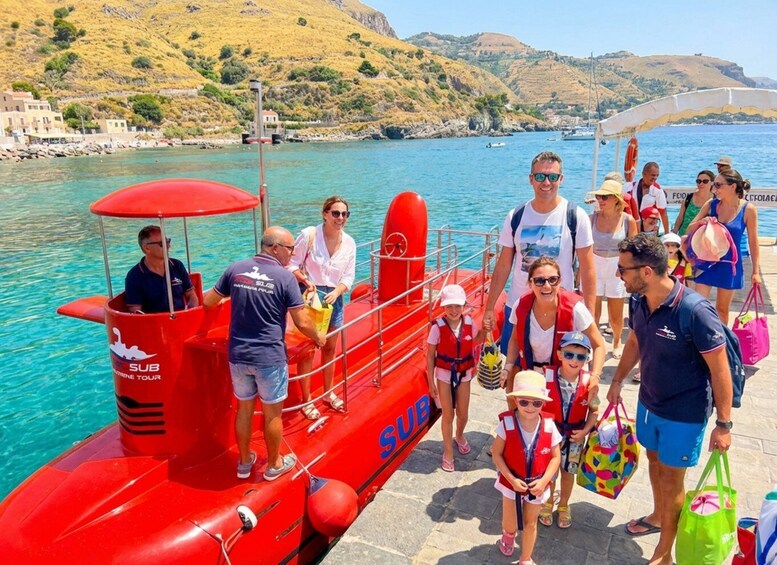 A group of people on a boat with a red 'SUB' sign, near a coastal area with clear blue water and hills in the background. 4