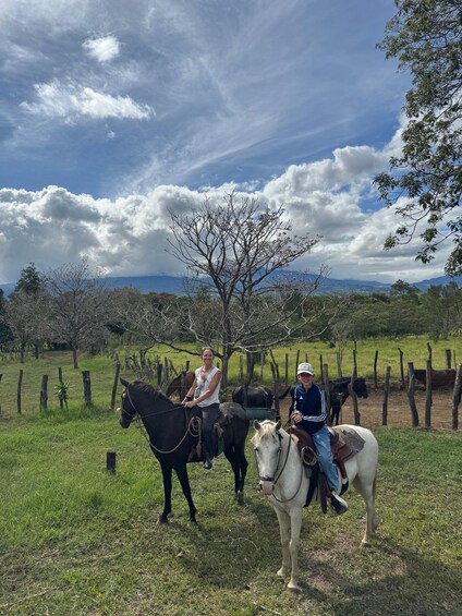Two people riding horses in a grassy field with a tree and a fence in the background. 2