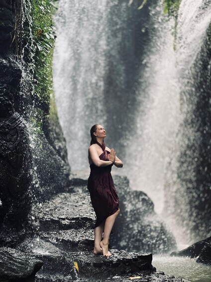 Una persona con un vestido color burdeos parada descalza sobre escalones rocosos con una cascada al fondo. 3