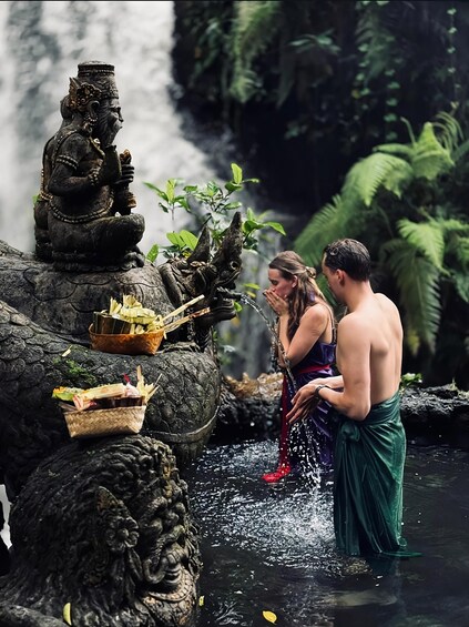 Una estatua de una figura sentada con ofrendas, una cascada y dos personas vestidas con atuendo tradicional junto al agua. 1