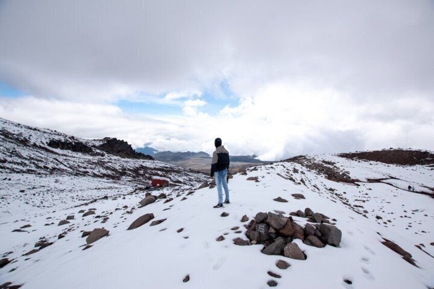 A person standing on a snow-covered mountain path with a rocky outcrop in the foreground and a cloudy sky above. 5
