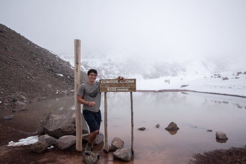 A person standing next to a signpost with 'CONDOR COGHA 5100 m' written on it, near a body of water and rocky terrain. 1