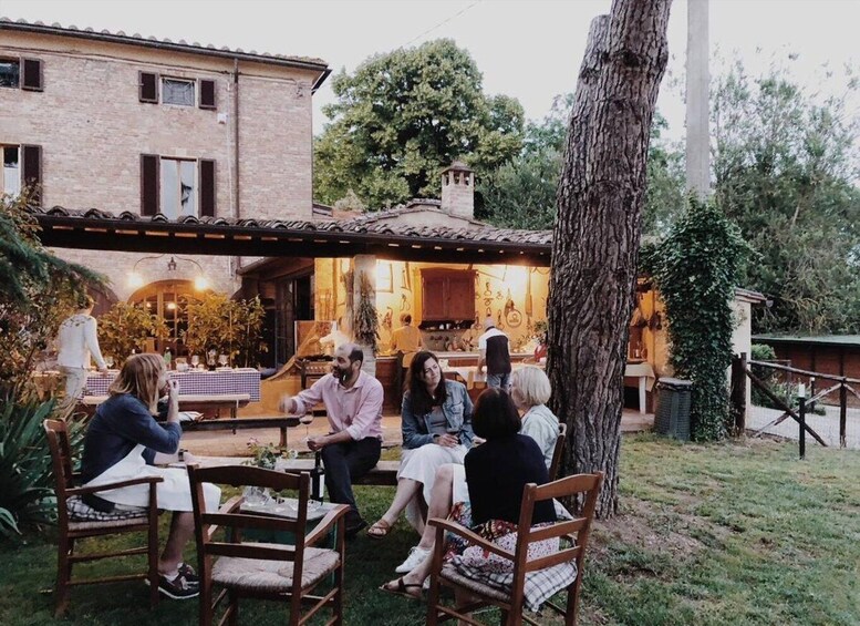 A group of people seated at an outdoor table in a rustic outdoor dining area with a brick building and trees in the background. 1