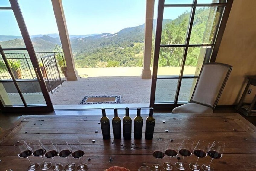 A wine tasting setup with bottles and glasses on a wooden table, overlooking a mountainous landscape. 1