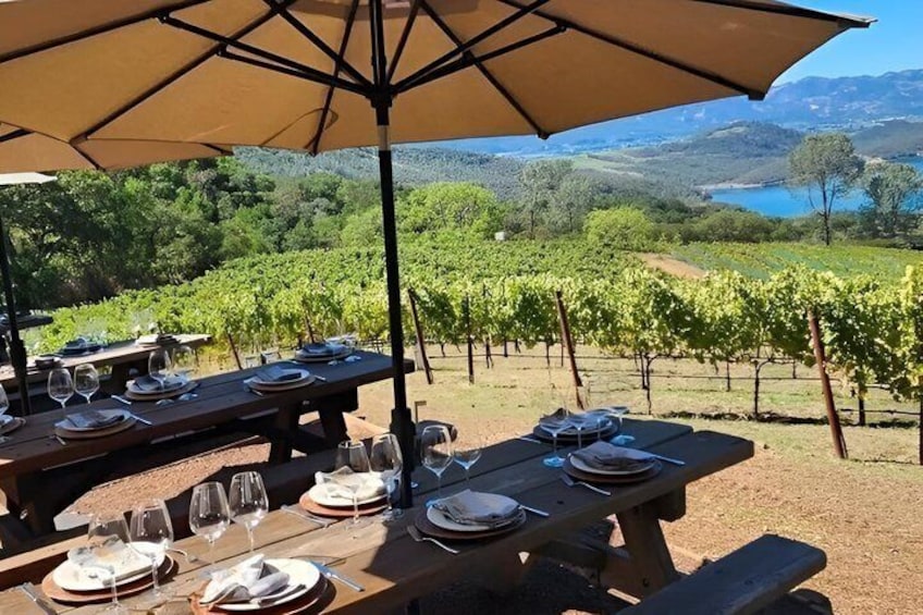 Outdoor dining tables set up with wine glasses and plates, overlooking a vineyard and a lake. 3