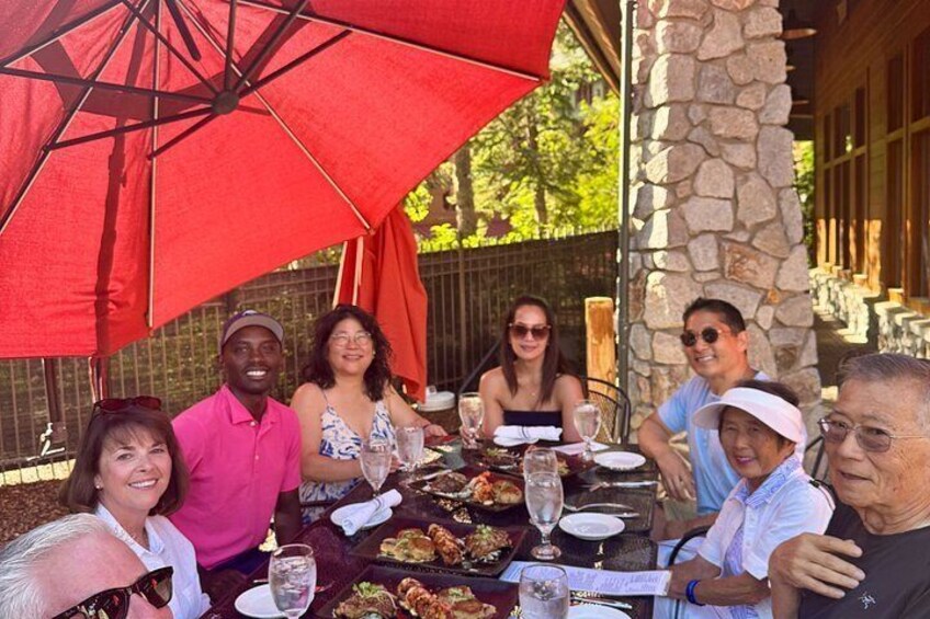 A group of people dining outdoors at a table with a red umbrella, stone pillar, and greenery in the background. 5