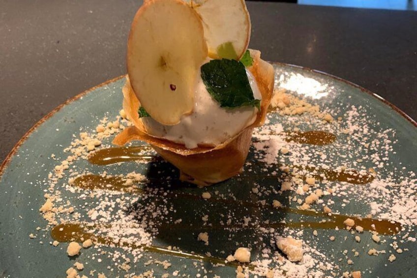 A dessert with a slice of apple, mint, and a creamy topping on a plate with powdered sugar and syrup. 5