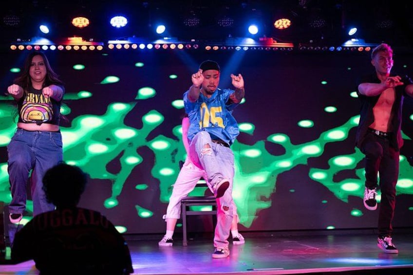 A stage with performers in casual attire, a green lighted backdrop, and a crowd in the foreground. 3