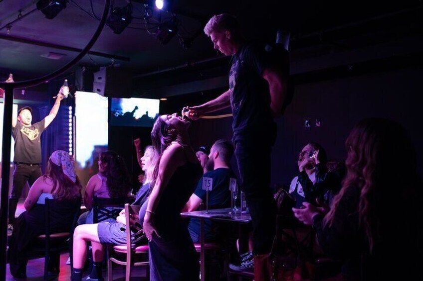 A person standing on a chair, possibly performing, in a dimly lit room with a crowd seated at tables. 4