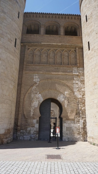 A historical stone archway with a person standing in front, a clear sky, and a paved ground. 2