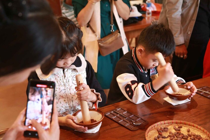 People grinding spices in bowls, a smartphone recording, and a tray of cocoa beans. 5