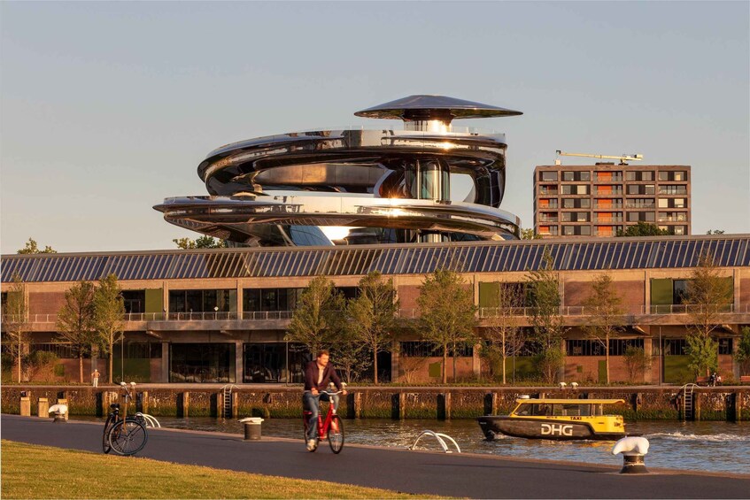 A modern building with a circular design, a cyclist on a path, and a boat with 'DHG' on the water. 1