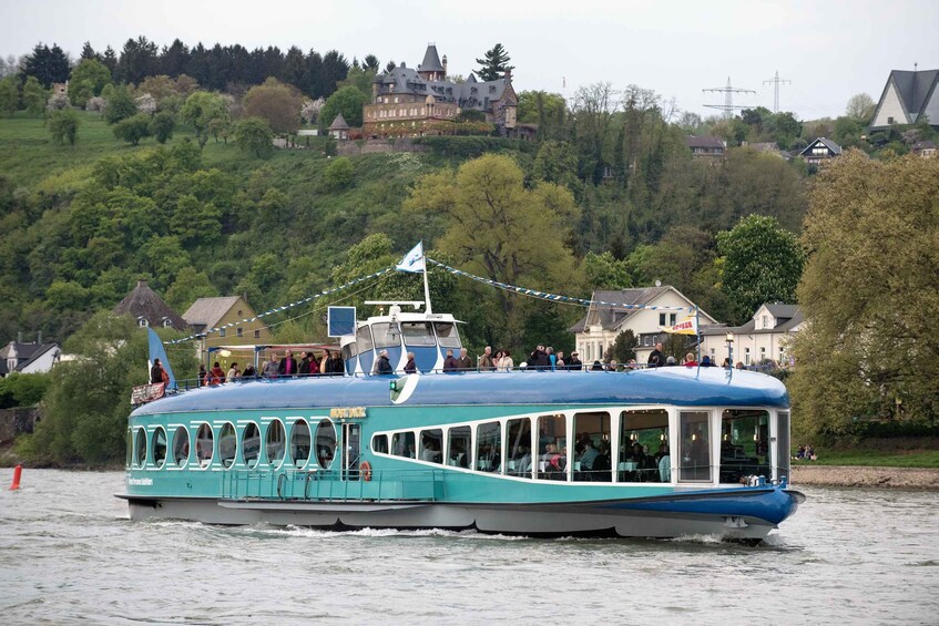 A blue river cruise boat with passengers, moving past a forested hillside with houses. 5
