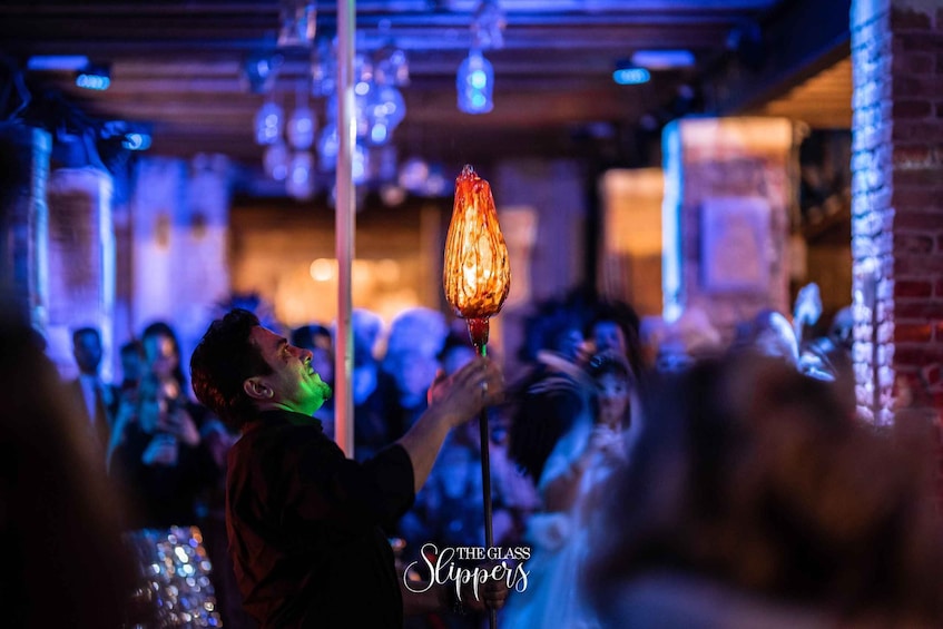 A person holding a lit glass with a flame, in a dimly lit indoor setting with brick walls and hanging lights. 2