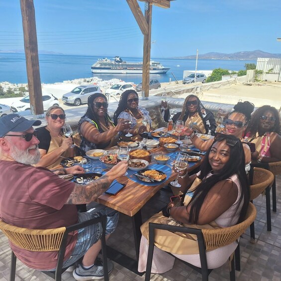 A group of people dining outdoors with a view of the sea and boats in the background. 4