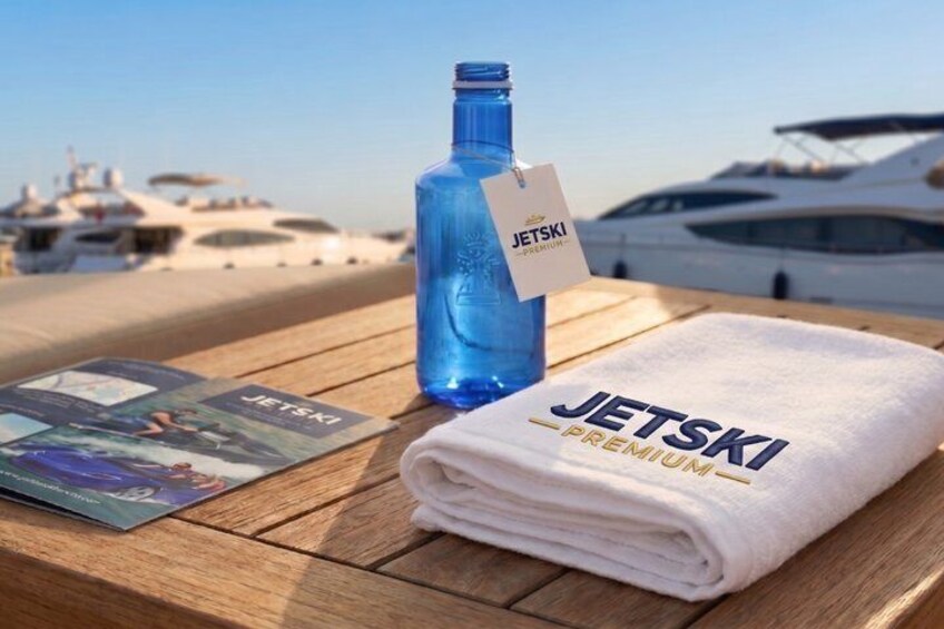 A folded towel with 'JETSKI PREMIUM' on it, a blue bottle, and a magazine on a wooden table with yachts in the background. 3