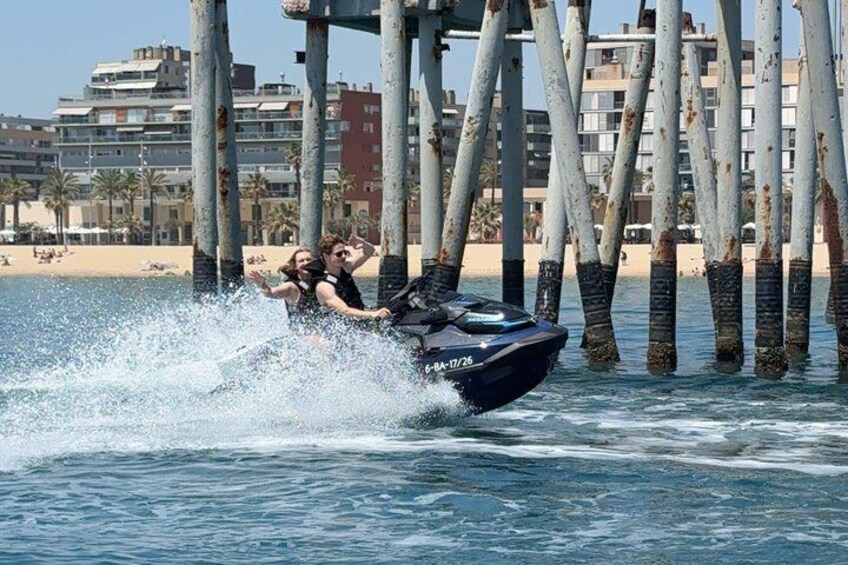 A jet ski with two people riding it, water splashing around, and a pier with buildings in the background. 2