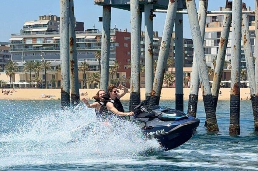 A jet ski with two people on it, water splashing around, a pier with trees, and a beach with buildings in the background. 5