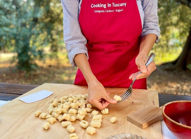 A person in a red apron labeled 'Cooking in Tuscany' preparing pasta on a wooden table outdoors. 5