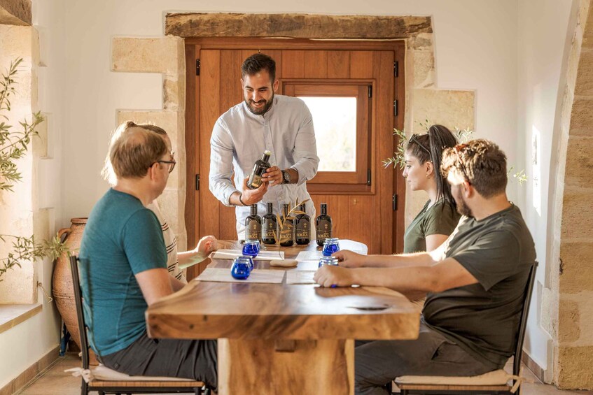 A group of people seated around a wooden table with bottles and glasses, in a room with stone walls and a wooden door. 1