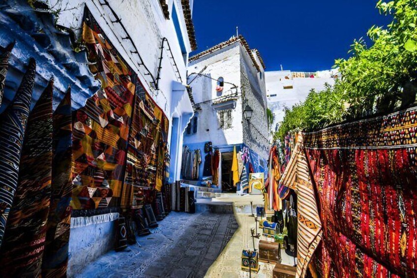 A narrow street with colorful textiles displayed outside shops, a white building with blue trim, and a clear blue sky. 5