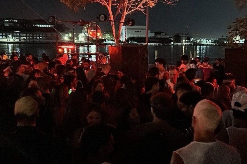 A crowded outdoor event by a waterfront with a stage and trees. 2