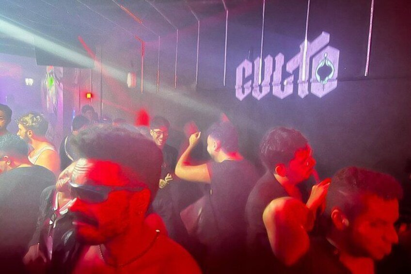 A crowded indoor venue with a neon sign reading 'CULTA' and people dancing. 5